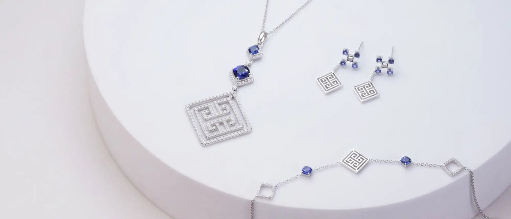 Silver necklace featuring a square greek key pendant adorned with blue sapphires and pavé diamonds, complemented by matching earrings and bracelet.